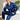 Man in blauwe satijnen pyjama met witte biezen, zittend in moderne kamer met plant.