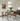Eetkamer met glazen tafel, houten stoelen, moderne hanglamp, groene plant, schilderij aan muur.