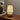 Beige tafellamp met linnen kap op houten tafel naast bloemen en boeken, sfeerverlichting.